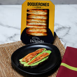 A package of Delgado Boquerones Al Picentón stands behind a black plate holding two slices of grilled bread topped with green avocado and cheese on a woven placemat, with a marble surface and folded red napkin in the background.