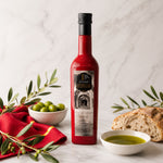 A bottle of Castillo de Canena Extra Virgin Olive Oil stands on a light marble surface, styled with a bowl of green olives, a slice of rustic bread, a bowl of olive oil, and olive branches.
