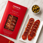Fermín Serrano Loin ham packaging in red, shown beside sliced cured ham on a white plate and a small bowl of green olives on a light marble surface.