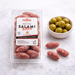 Packaged salami bites with a bowl of green olives on a marble surface