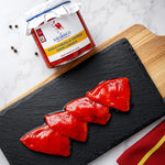 A glass jar of Navarrico whole roasted piquillopeppers from Lodosa sits on a marble surface next to a wooden board and black slate platter holding four glossy red pepper slices.