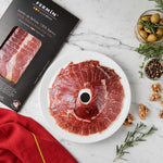 Sliced cured ham arranged on a white plate with a black Fermin & Cía Modelo package beside it, styled on a white marble surface with olives, rosemary, peppercorns, walnuts, and a red cloth.
