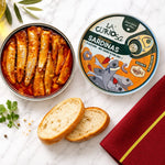 An open round tin of La Curiosa Sardinés Sardinas with salsa brava, shown on a white marble surface with bread slices, parsley, olives, and a red napkin.