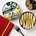 A top-down styled food presentation on a white marble surface. The main product is a round tin of La Curiosa Sardinas in truffle olive oil, with the lid shown on the left and the open tin filled with cooked sardines on the right.