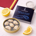 Oysters in a tin with a box labeled 'La Mar de Tazones' on a white surface with lemon slices and salt.