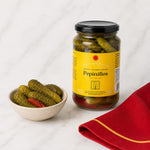 A jar of Pepinillos pickles with a yellow label and black lid stands on a white marble surface next to a small bowl of whole pickles and a folded red cloth with a gold stripe.