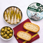 Canned sardines, green olives, and bread slices on a white surface with 'La Curiosa' packaging.