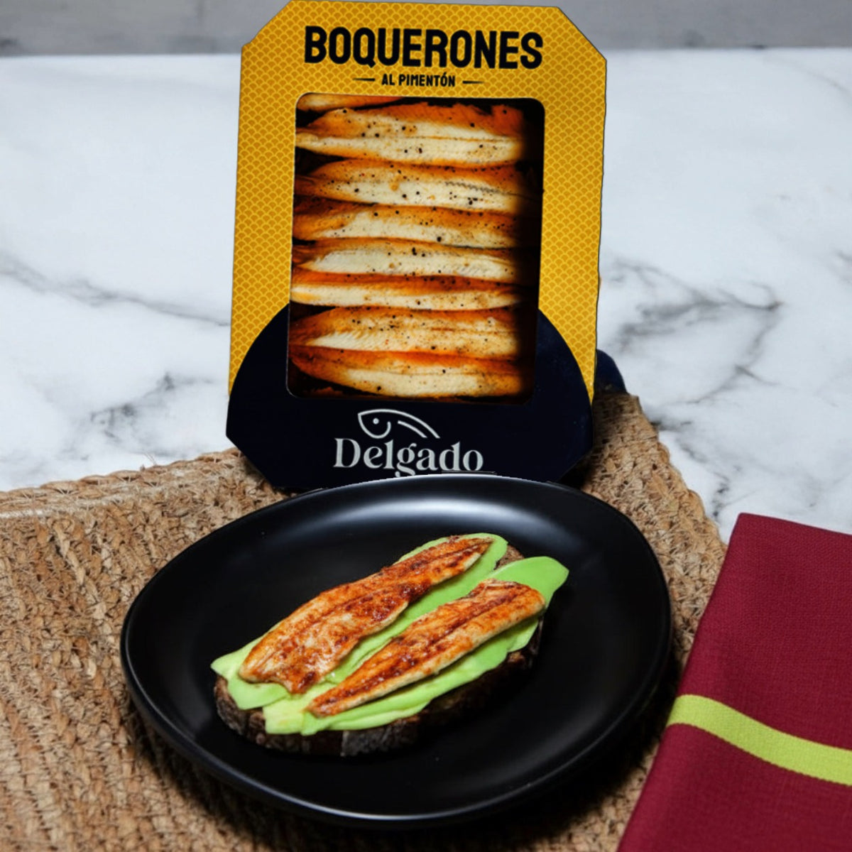 A package of Delgado Boquerones Al Picentón stands behind a black plate holding two slices of grilled bread topped with green avocado and cheese on a woven placemat, with a marble surface and folded red napkin in the background.