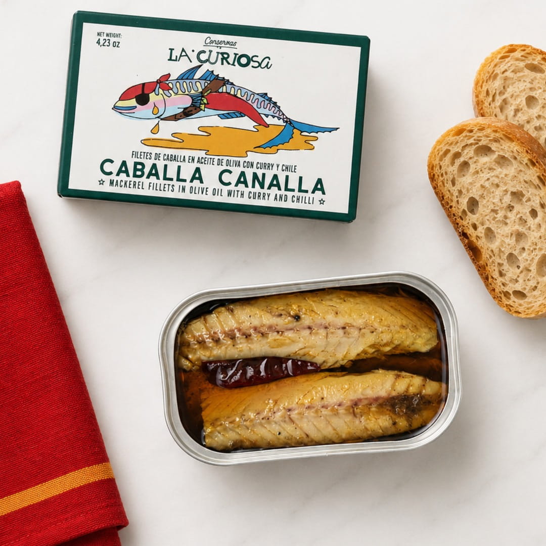 A tin of La Curiosa Caballa Canalla mackerel fillets in olive oil with curry and chili, shown next to a red cloth napkin and two slices of bread on a white surface.