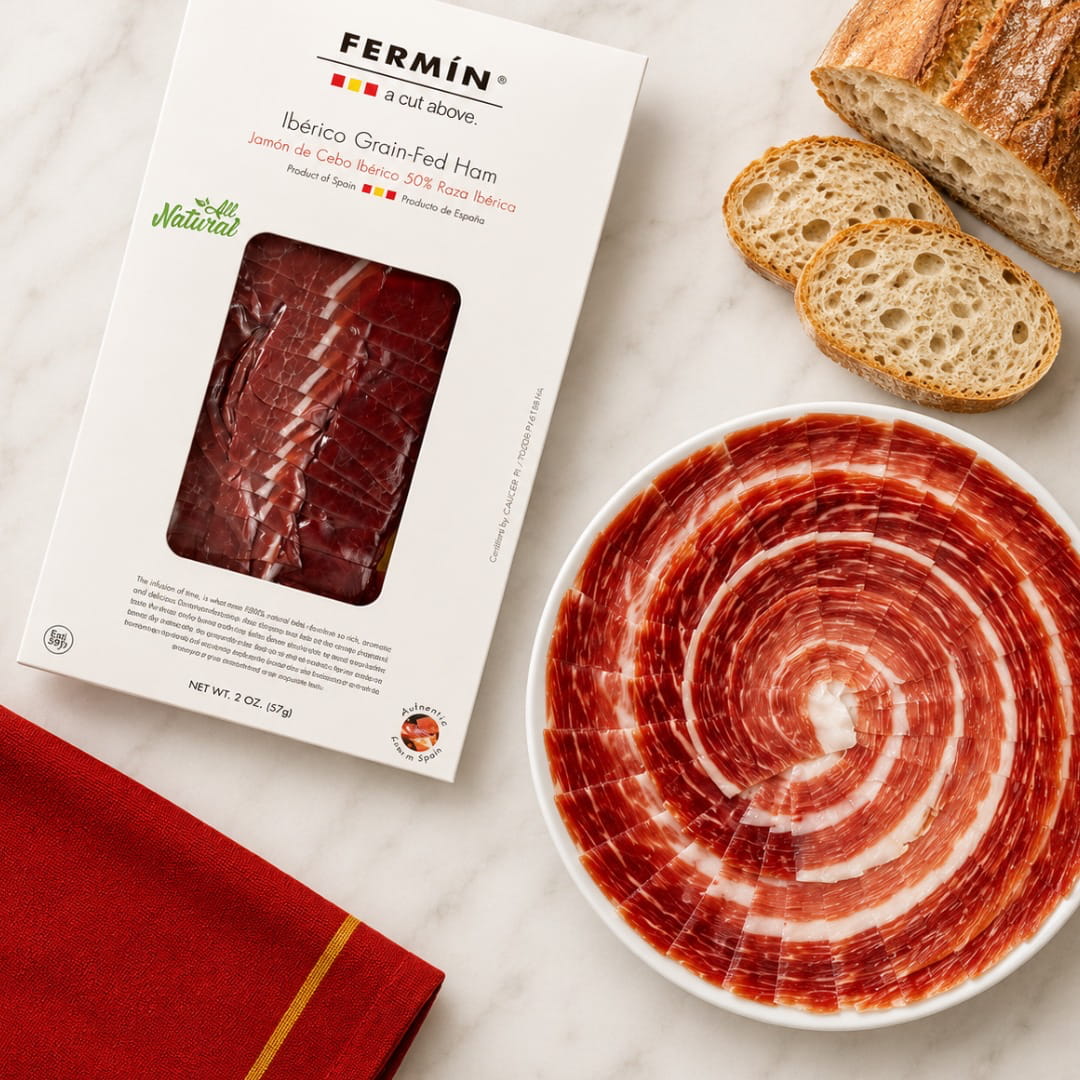 A package of Fermin Ibérico Grain-Fed Ham is shown on a white marble surface next to a sliced loaf of bread and a white plate filled with thinly sliced ham arranged in a spiral pattern. A red cloth with a gold stripe is visible in the lower left corner.
