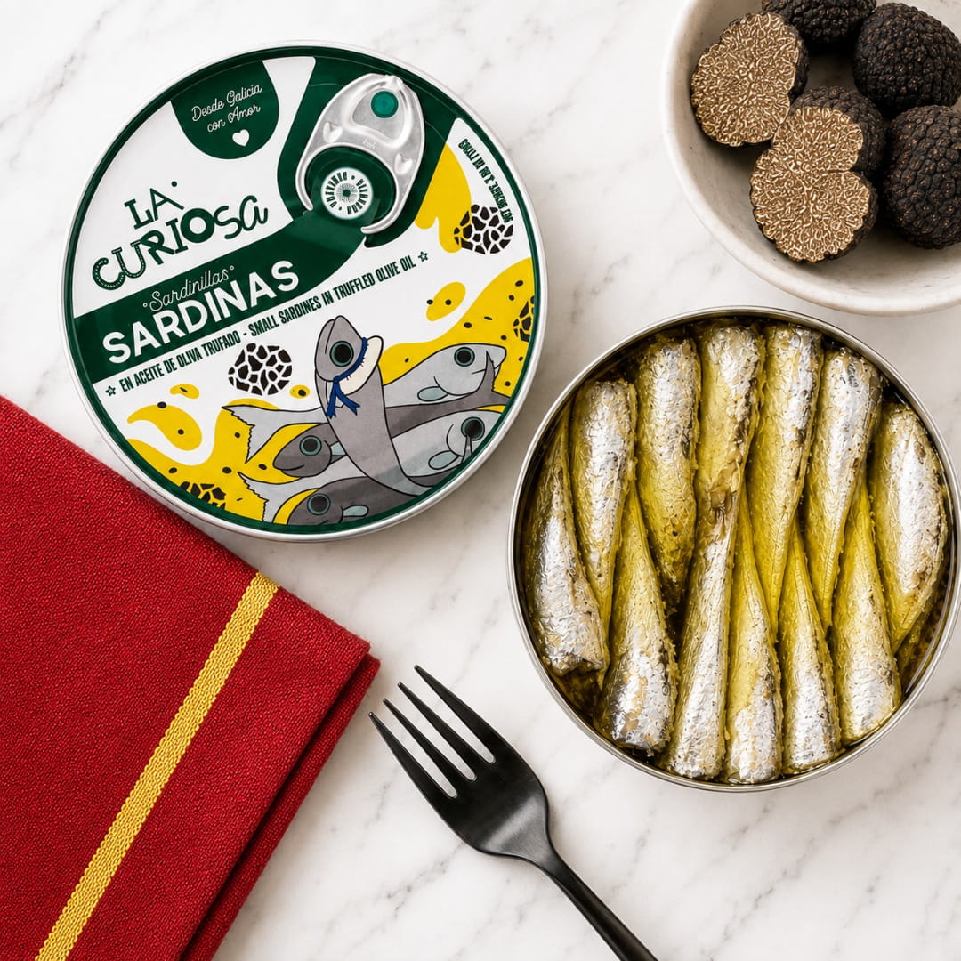 A top-down styled food presentation on a white marble surface. The main product is a round tin of La Curiosa Sardinas in truffle olive oil, with the lid shown on the left and the open tin filled with cooked sardines on the right.