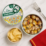 A top-down styled flat lay of La Curiosa Zamburinas on a white marble surface, featuring an open can of cooked scallops, a closed can with its illustrated label, a small bowl of potato chips, and a fork on a red napkin.