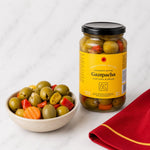A jar of Gazpacha seasoned olives with red peppers and carrots stands next to a small bowl filled with sliced olives, topped with a black lid, on a white marble surface with a red napkin.