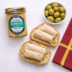 Jar of Bonito del Norte tuna, bread slices with tuna, and a bowl of green olives on a white background