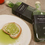 Two bottles of Oli del Reig olive oil with a piece of bread spread with green olive spread on a plate.
