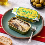 Can of sardines with 'Nuri' packaging on a plate with bread and olives.