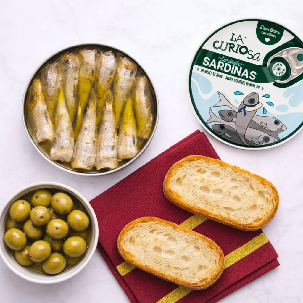 Canned sardines, green olives, and bread slices on a white surface with 'La Curiosa' packaging.