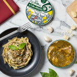 Pasta dish with pesto, a bowl of soup, and a container of La Curiosa Atun de Pesto on a wooden table.